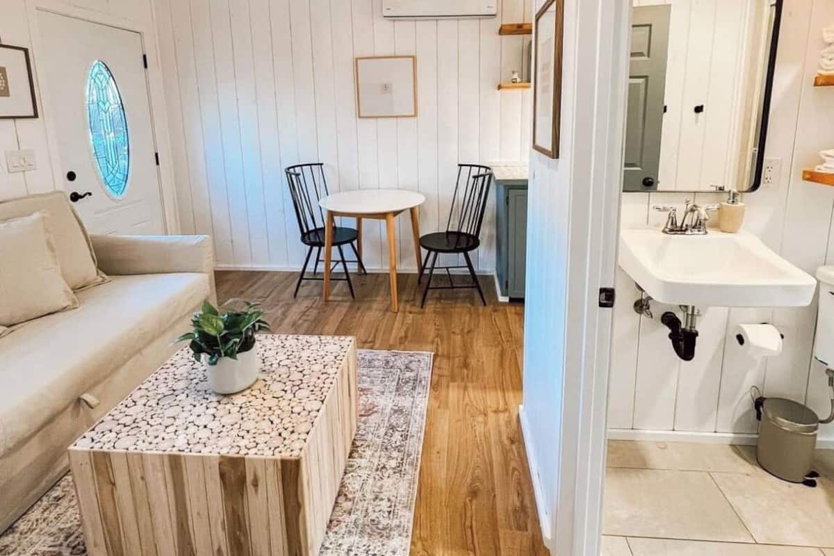 A cozy room with a wooden ceiling, white walls, a round table with chairs, a sofa, a coffee table with plants, and a bathroom with a sink and mirror.