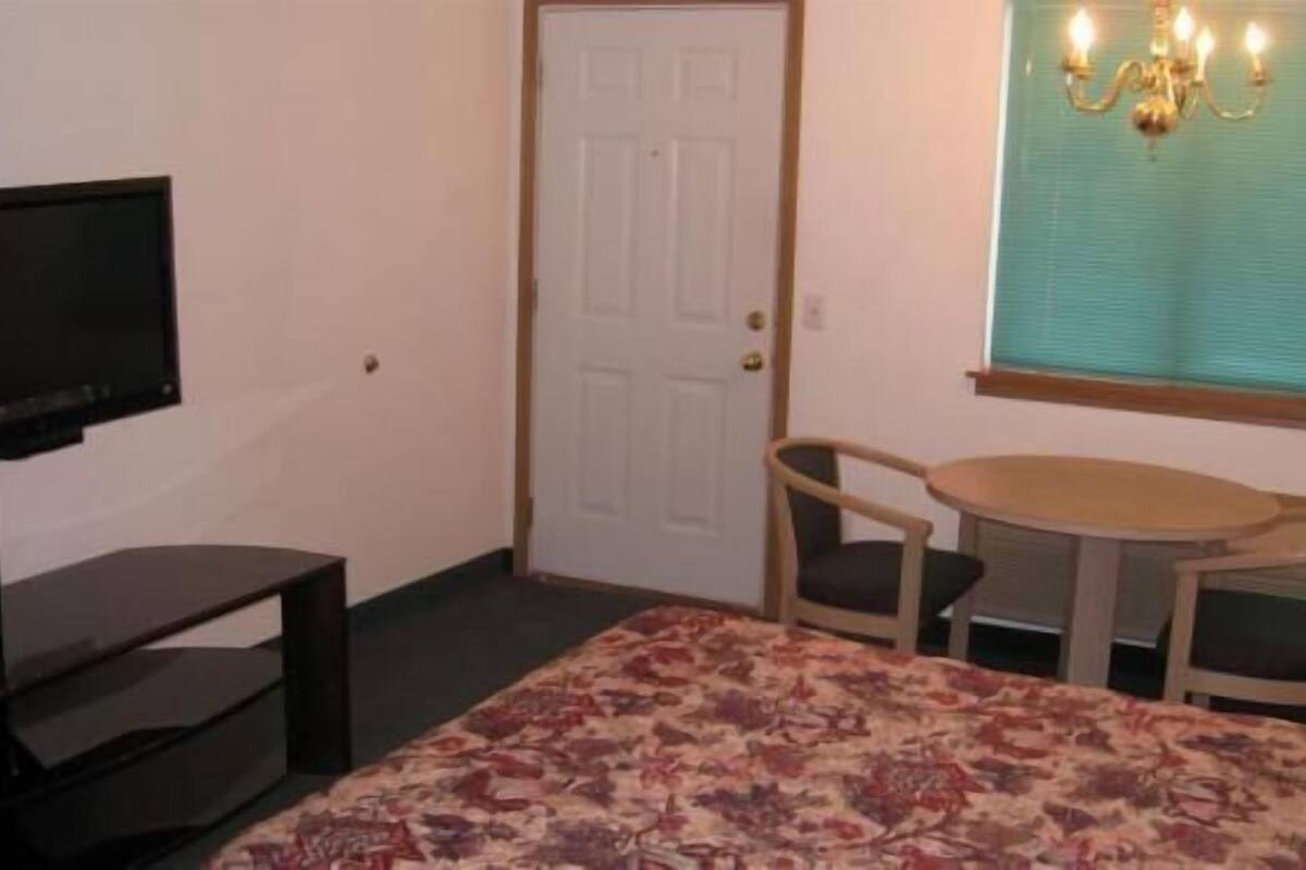 A room with a television on a stand, a bed with a floral bedspread, a small table with chairs, and a window with a curtain and a chandelier.