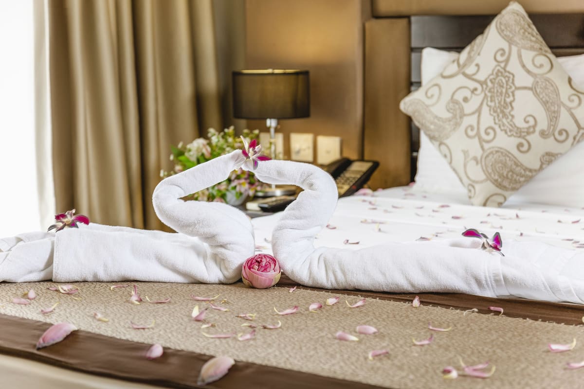 A neatly made bed with towels and flowers, a bedside lamp, and a telephone on a wooden nightstand.