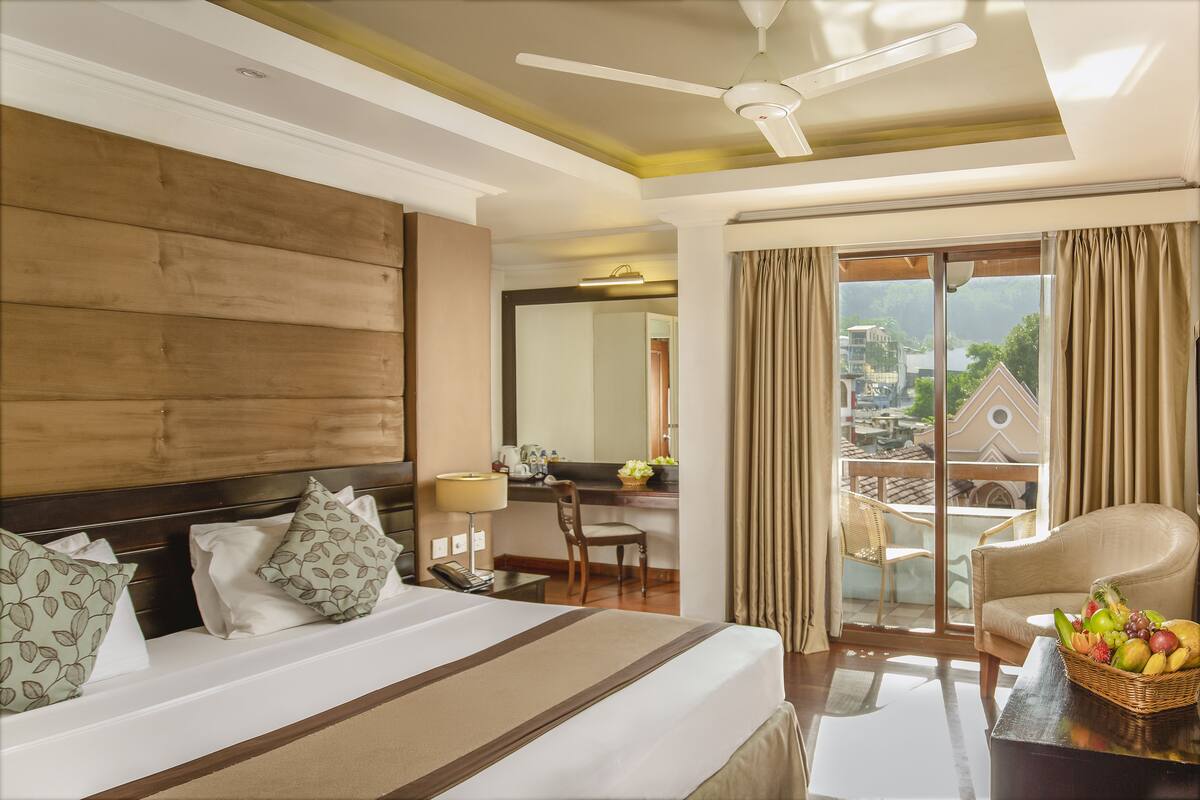 A hotel room with a large bed, a desk, a chair, a balcony with curtains, and a ceiling fan.