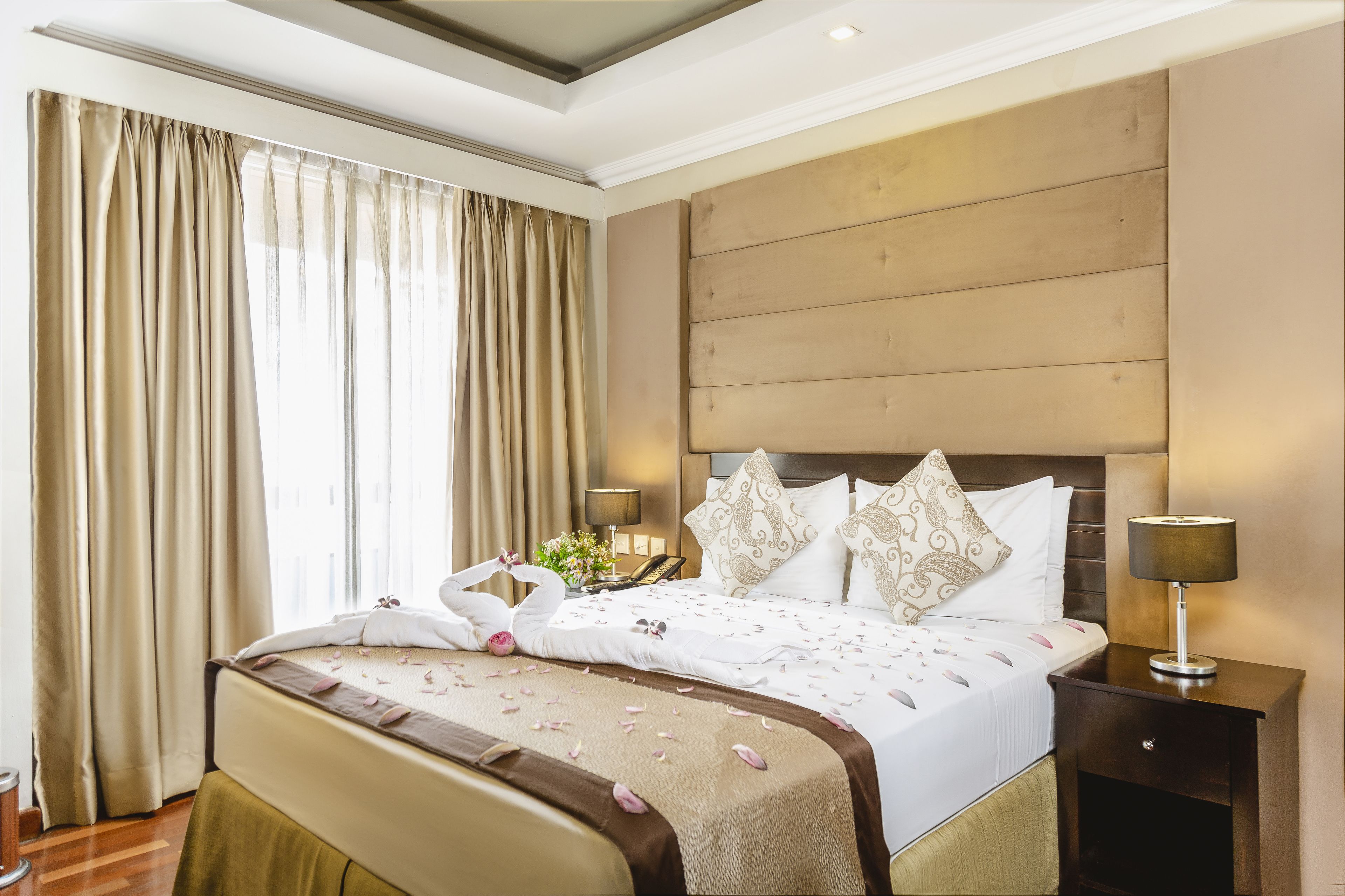 A hotel room with a large bed, a wooden headboard, a nightstand with a lamp, and a window with curtains.