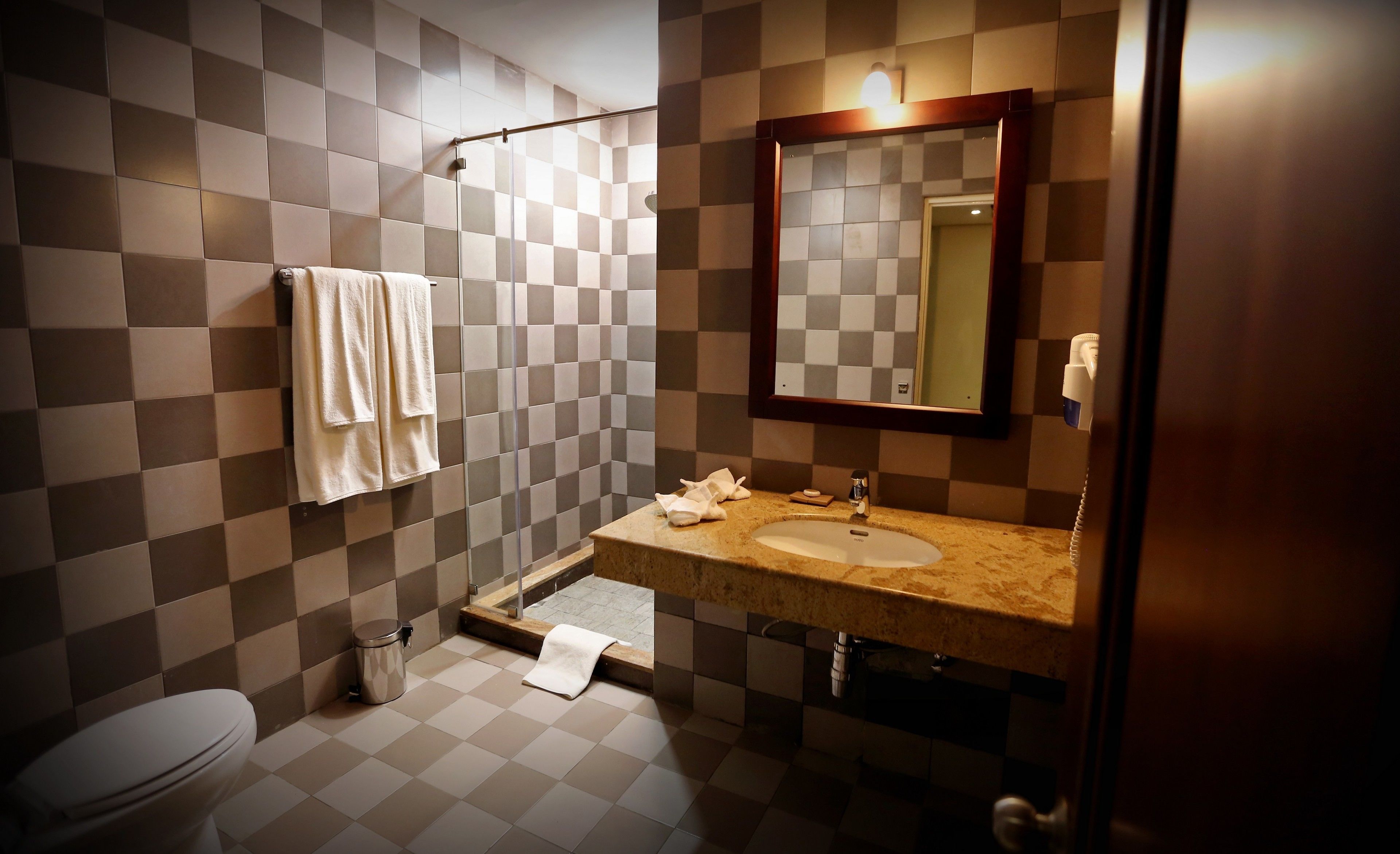 A bathroom with a checkered tile wall, a sink with a mirror, a toilet, and a trash can.