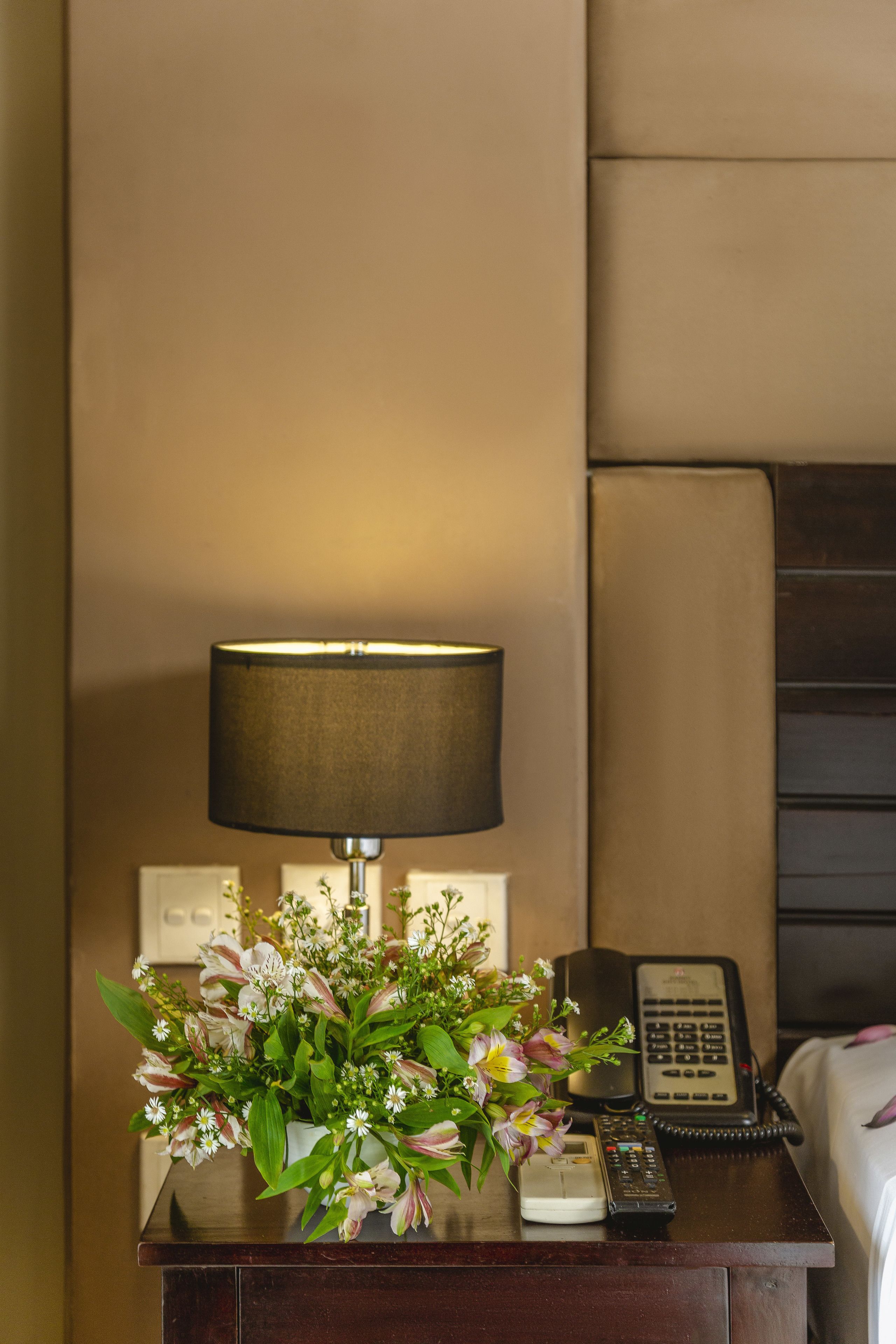 A wooden nightstand with a lamp, a telephone, and a floral arrangement.