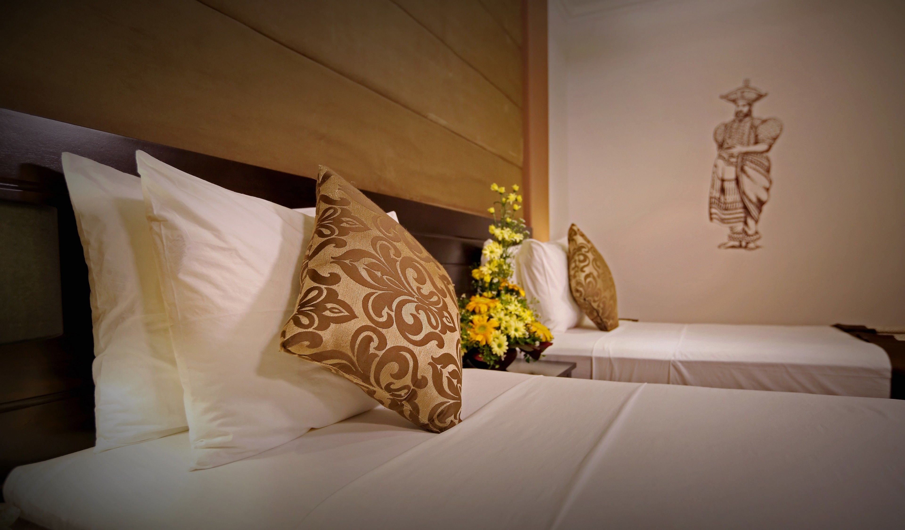 Dos camas con sábanas blancas y almohadas estampadas, una obra de arte en la pared y un arreglo floral.
