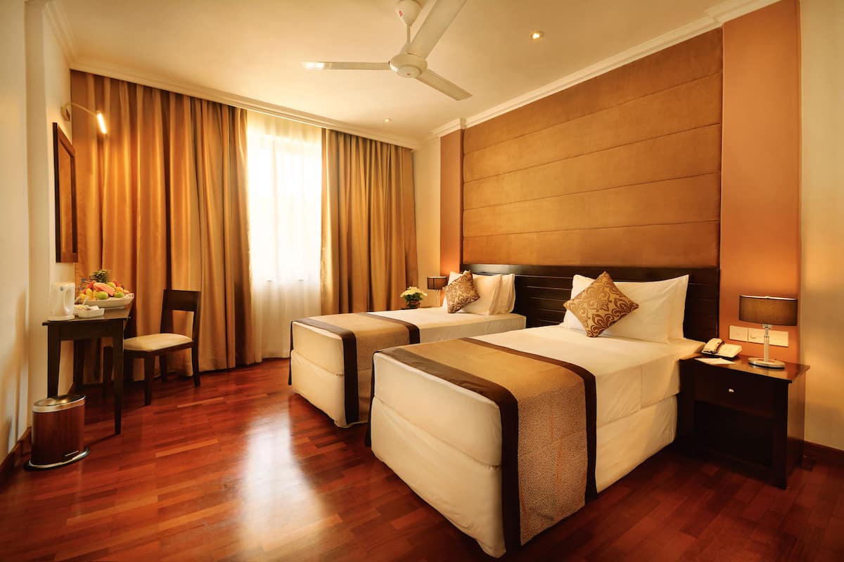 A hotel room with two beds, a desk, a chair, a trash bin, and a nightstand with a lamp.
