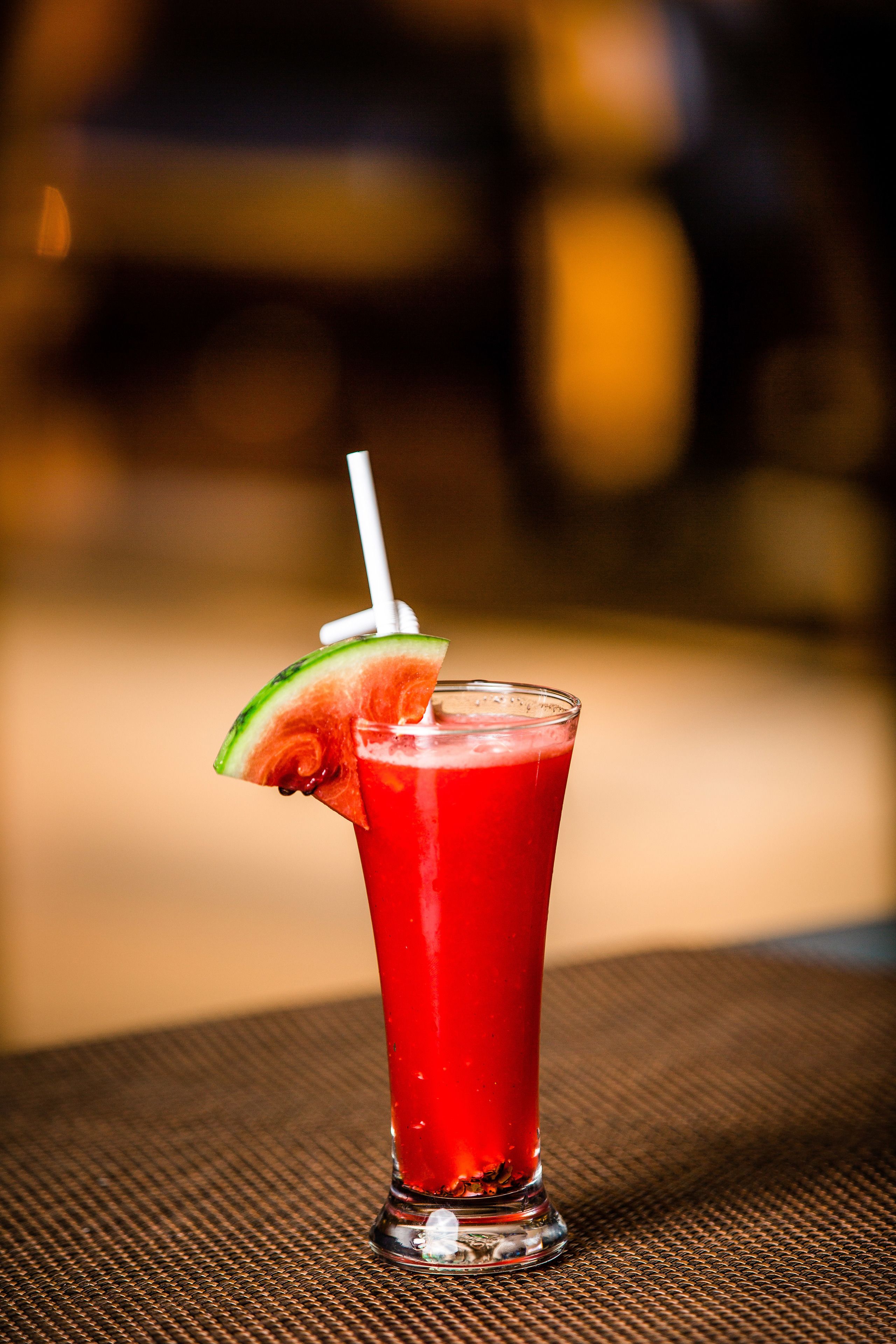 A tall glass with a red beverage, a slice of watermelon, and a straw on a textured surface.