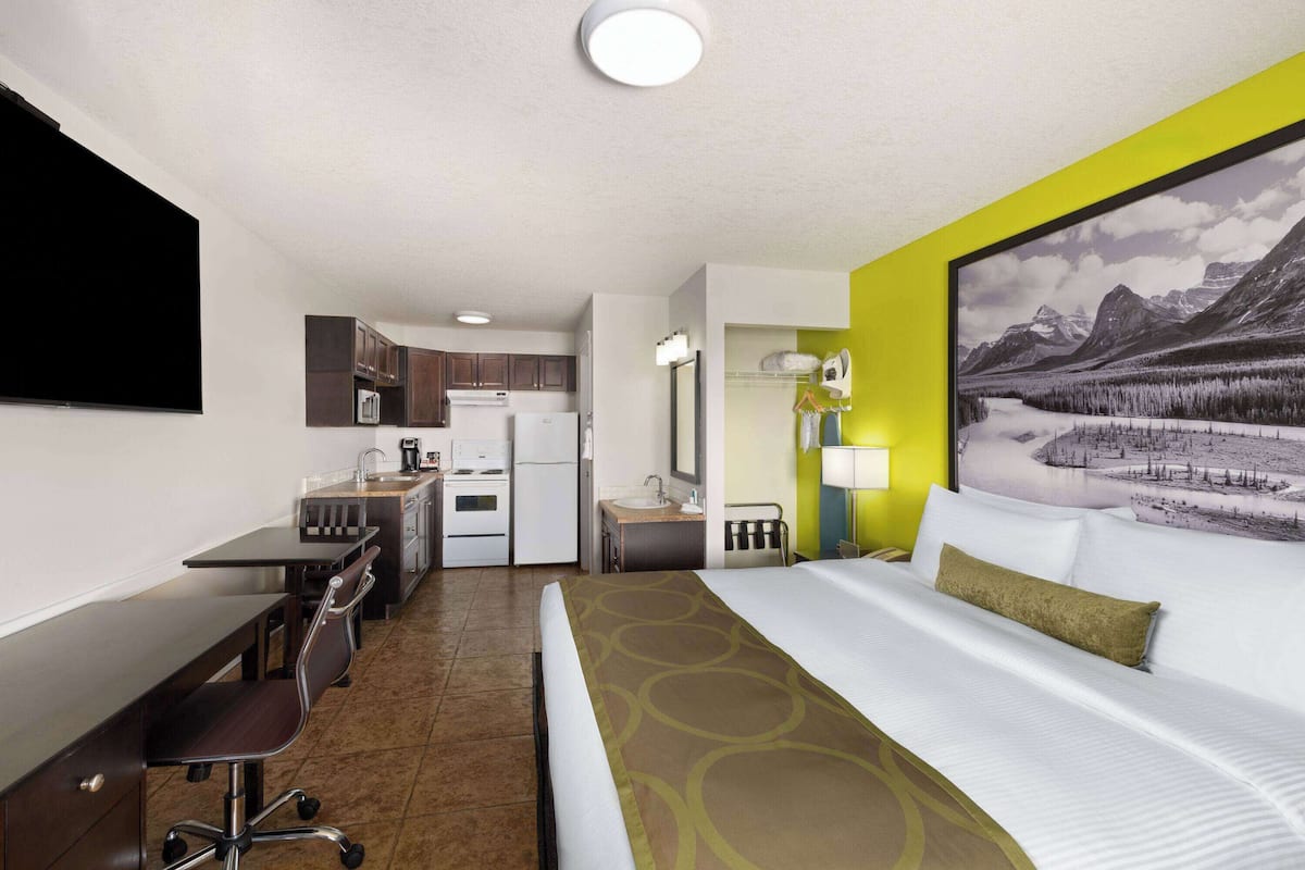 A hotel room with a bed, desk, chair, kitchenette, and a large window with a mountain view.