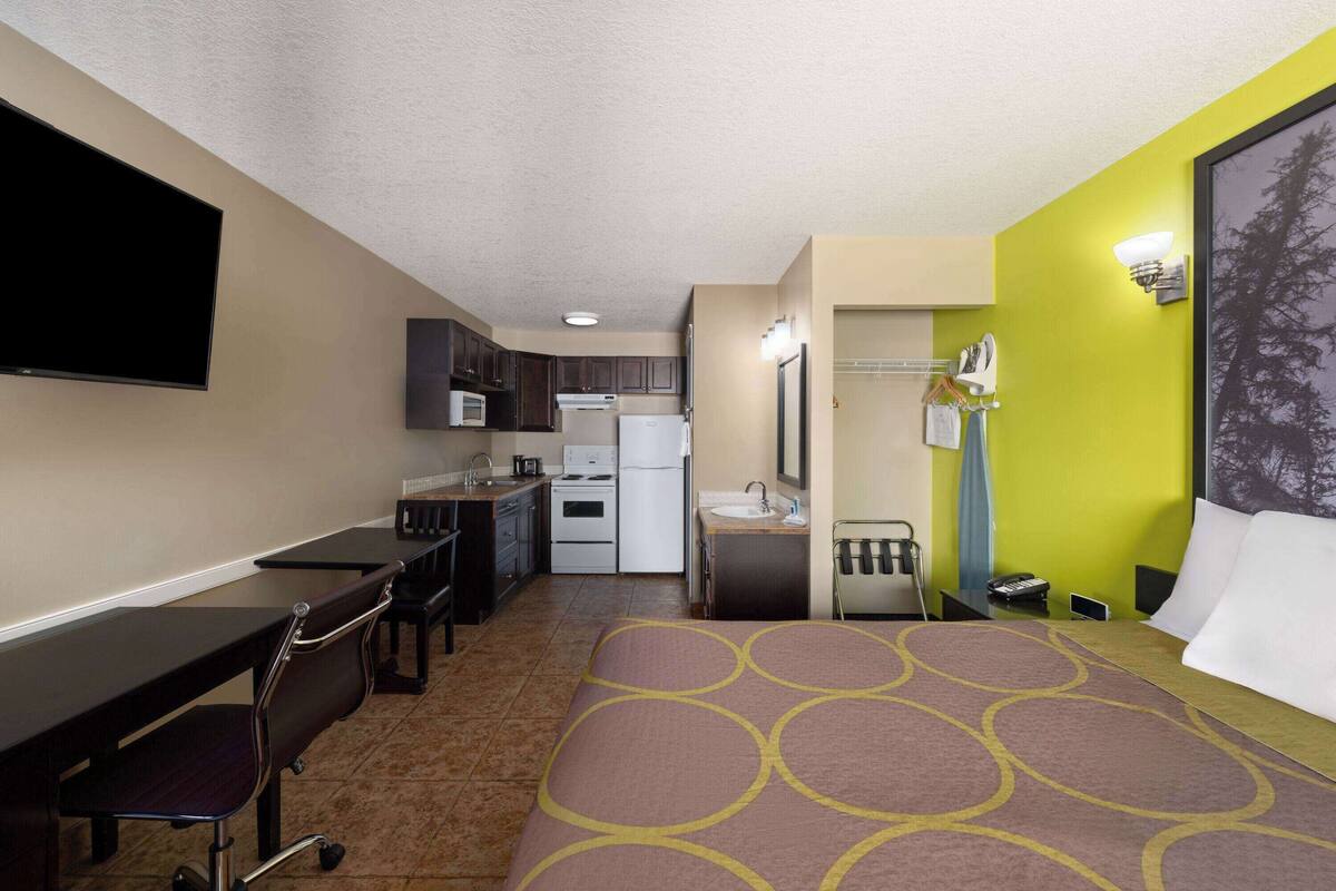 A compact hotel room with a kitchenette, a bed with a patterned bedspread, a desk with a chair, and a TV mounted on the wall.