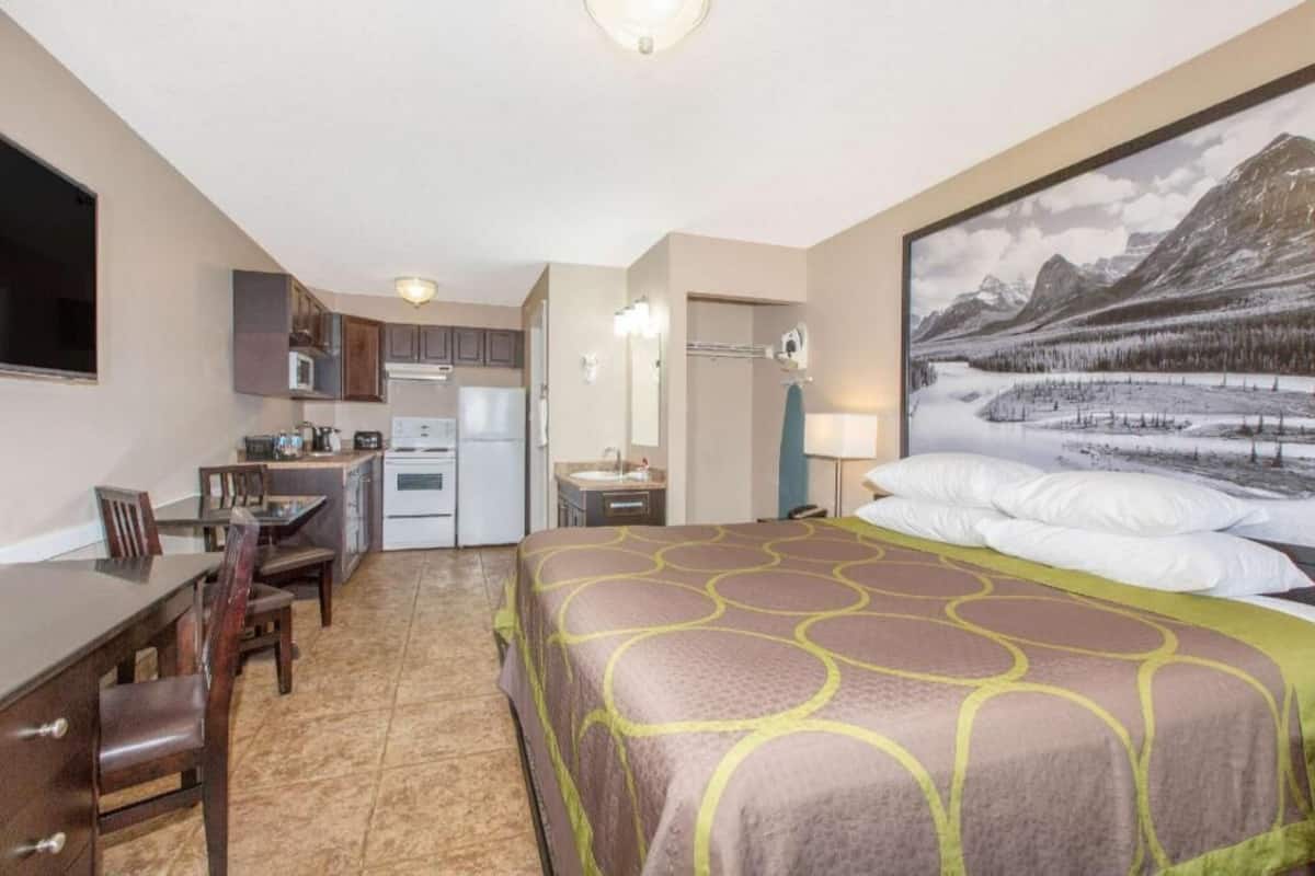 A hotel room with a bed, a desk, a kitchenette, and a large window with a mountain view.