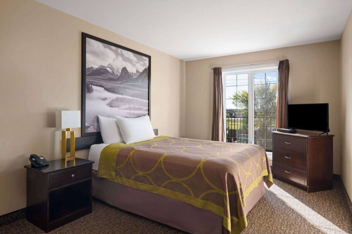 A hotel room with a bed, a nightstand with a phone, a dresser, a television, and a large window with curtains.
