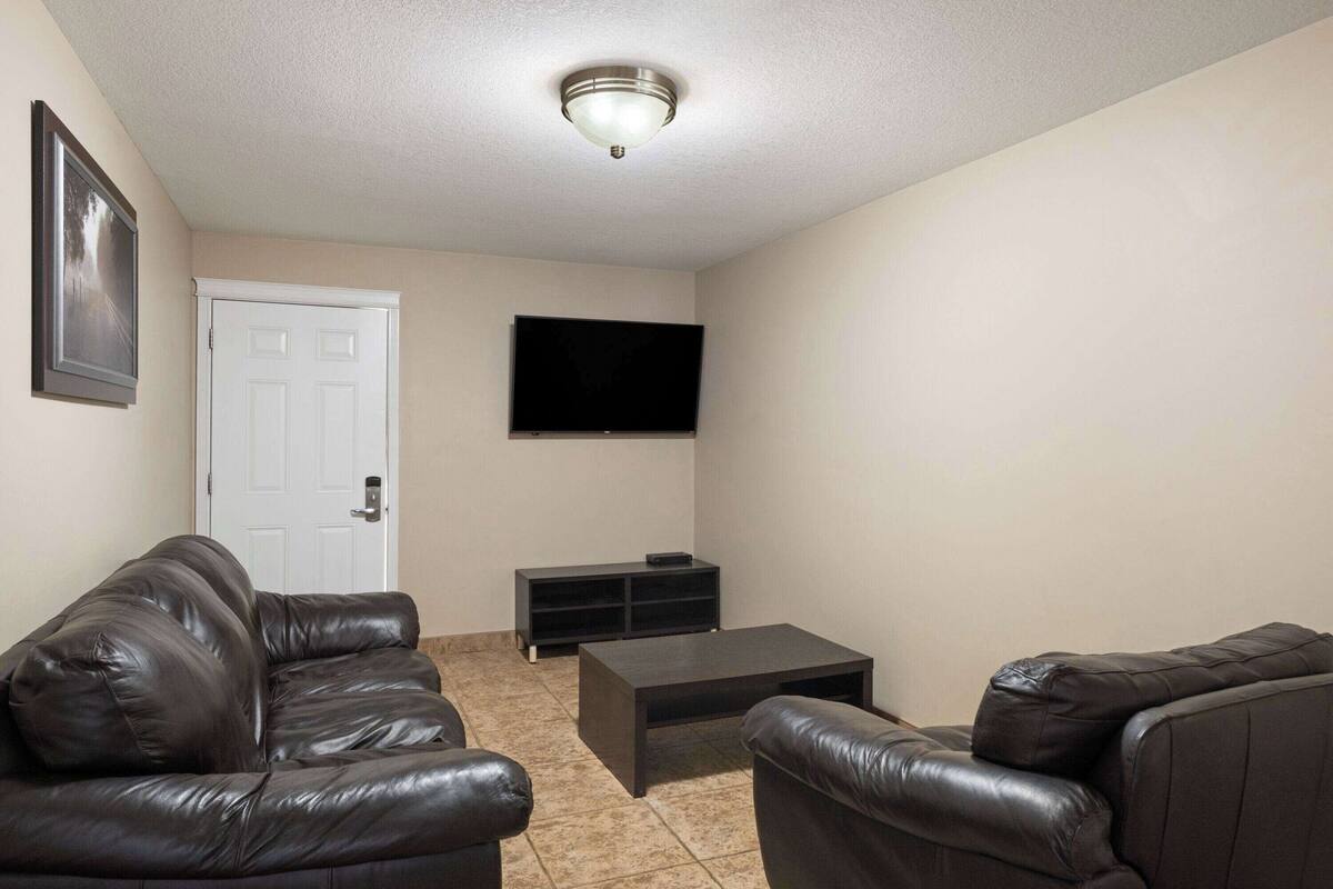 A living room with a flat-screen TV mounted on the wall, a dark brown leather sofa, a small coffee table, and a white door.
