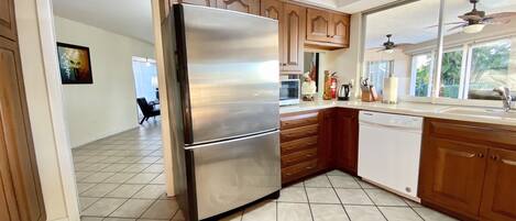 A kitchen with a stainless steel refrigerator, wooden cabinets, a dishwasher, and a window with a view of greenery.