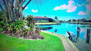 A swimming pool with a grassy area and palm trees, a dock with chairs, and a building in the background.