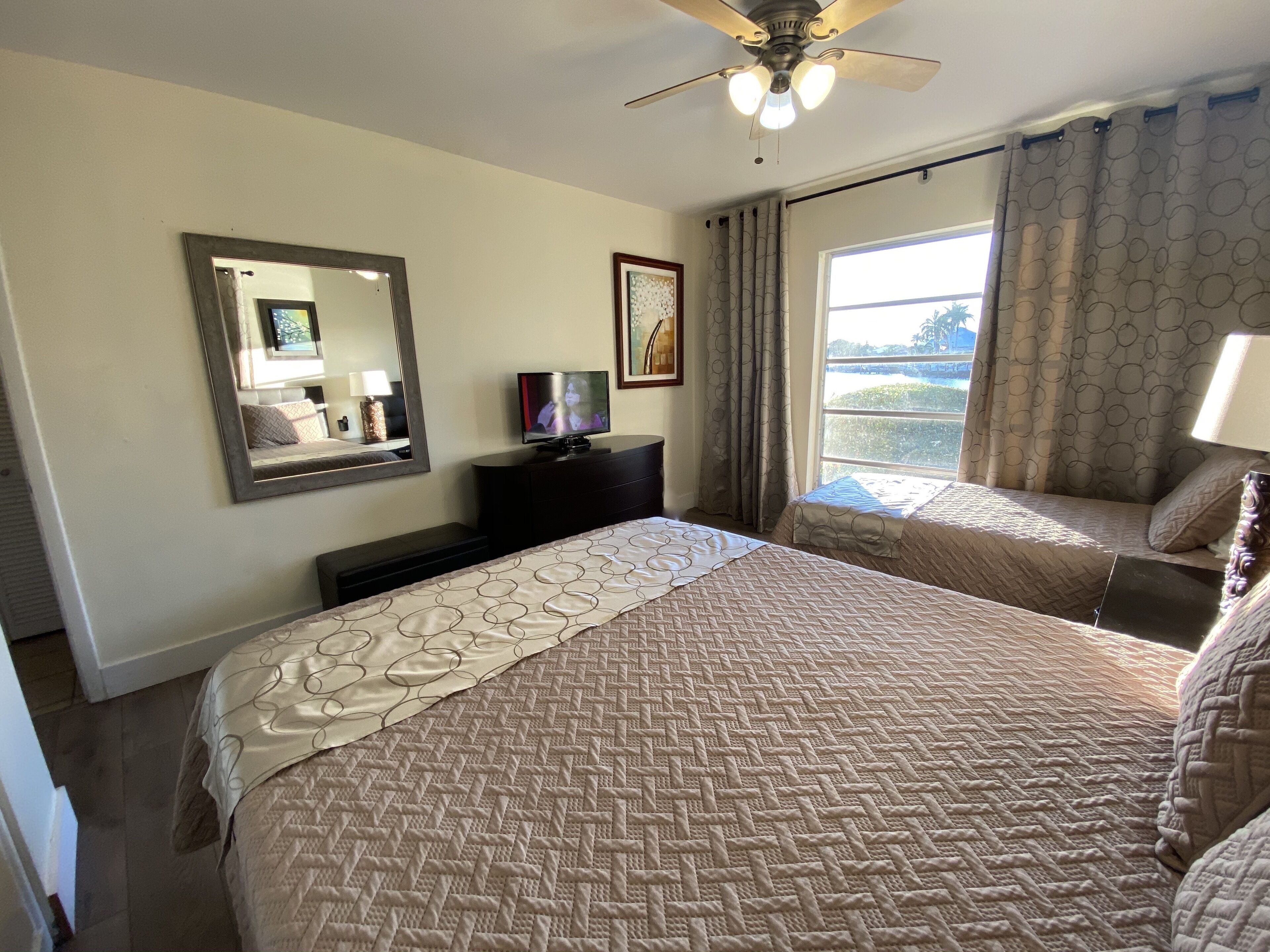 A hotel room with a large bed, a ceiling fan, a TV, a dresser, a window with curtains, and a painting on the wall.