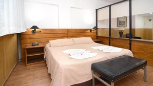 Een hotelkamer met een houten hoofdeinde, een bed met een beige deken, een zwarte bankje, een nachtkastje en een spiegel.