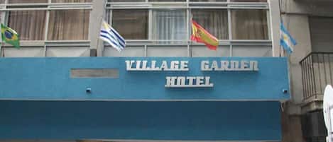 The exterior of the Village Garden Hotel with a blue awning and flags.