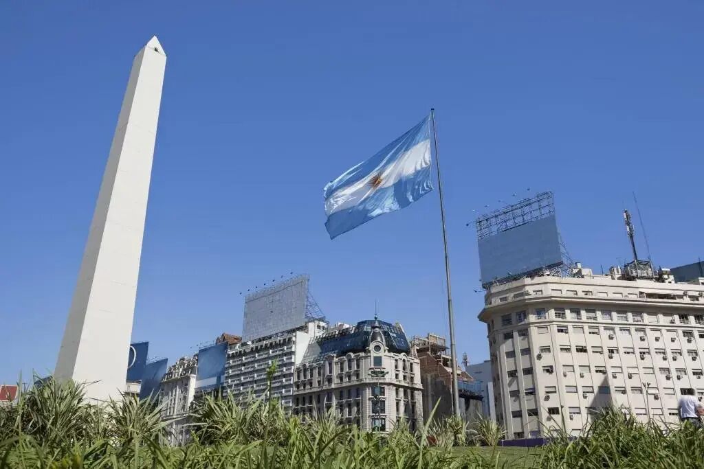 Un obelisco blanco y alto, una bandera y un edificio con cúpula y varias ventanas.