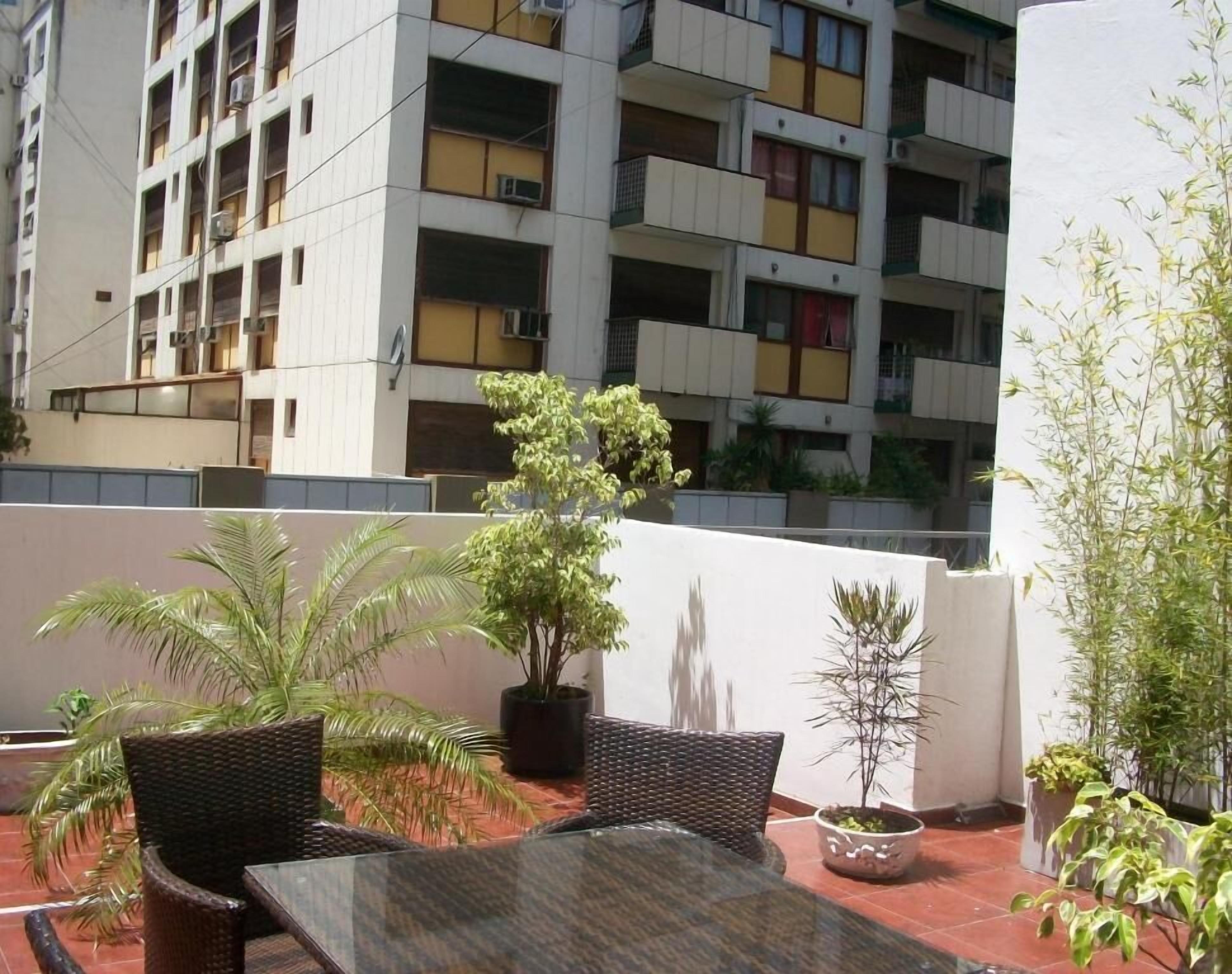 Un balcón con mobiliario de mimbre, plantas en macetas y vista a un edificio de varios pisos con balcones.