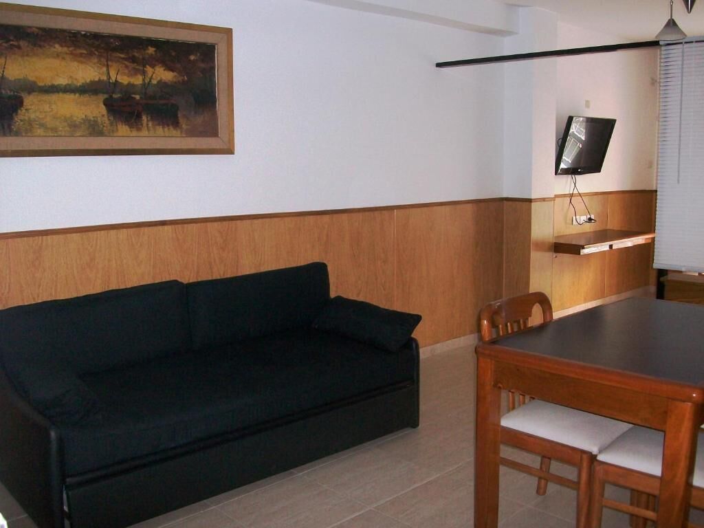 Et værelse med en sort sofa, et træspisebord med stole, et fjernsyn monteret på væggen og et billede over sofaen.