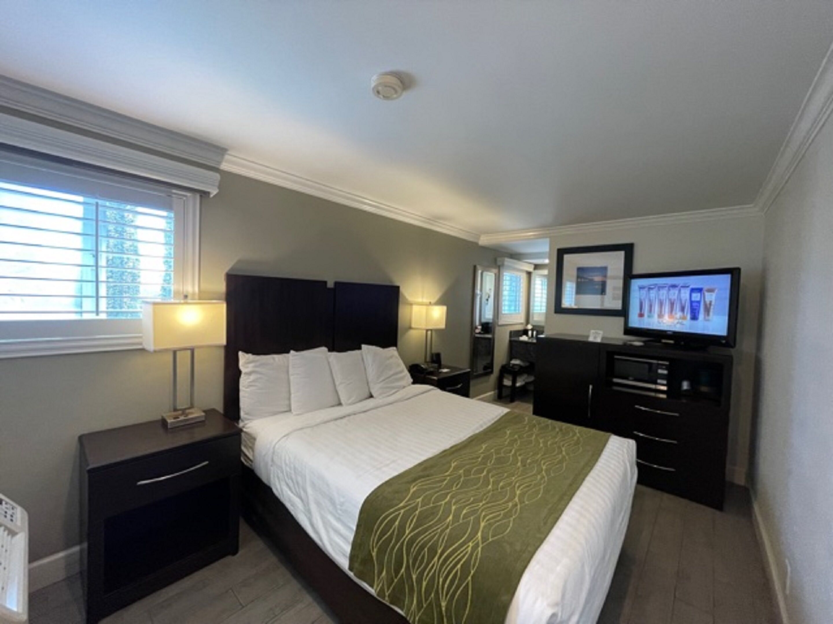 A hotel room with a bed, bedside tables, a desk, a television, and a window with blinds.