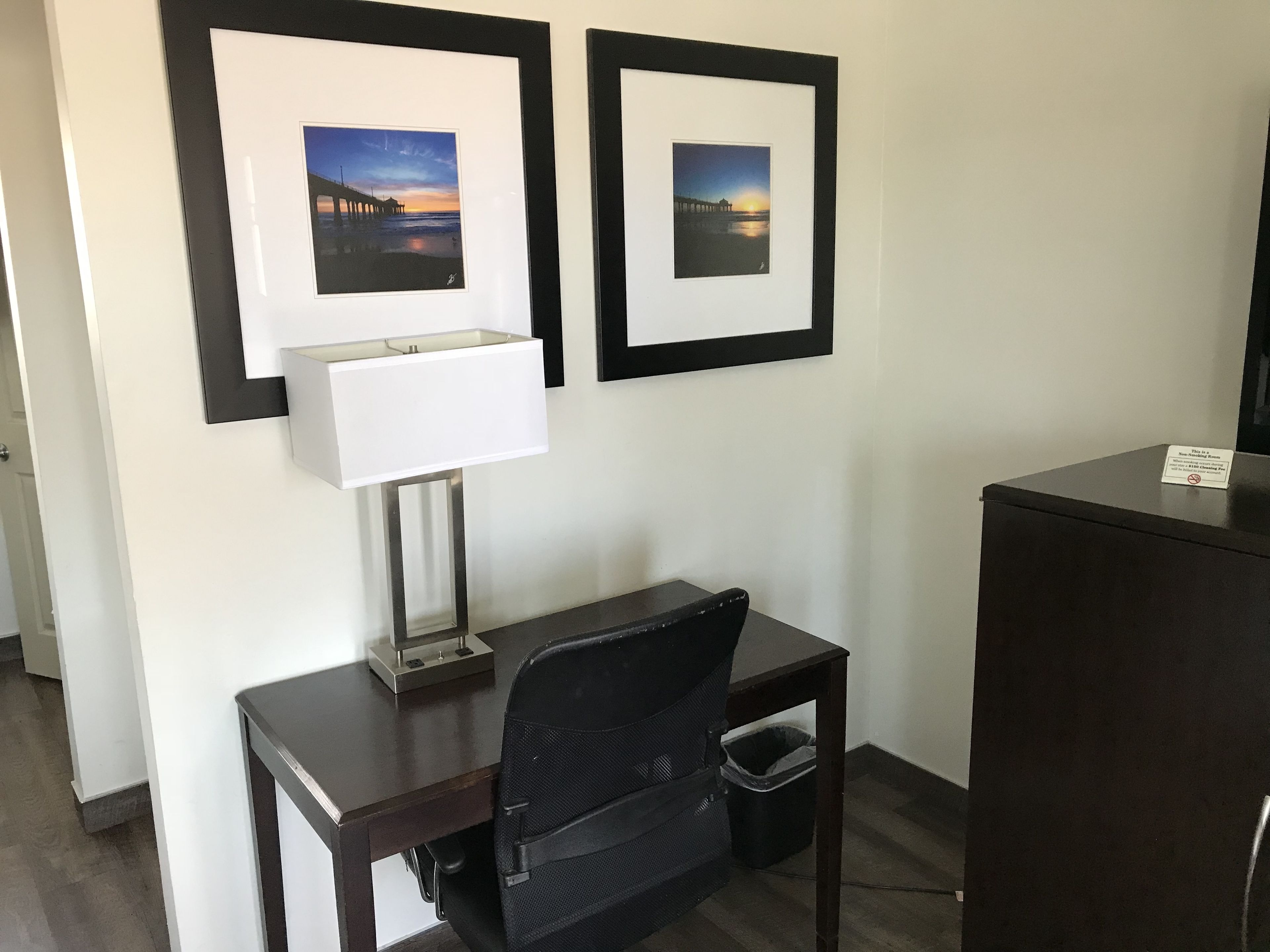 A hotel room with a desk, a chair, a lamp, a trash bin, and two framed pictures on the wall.