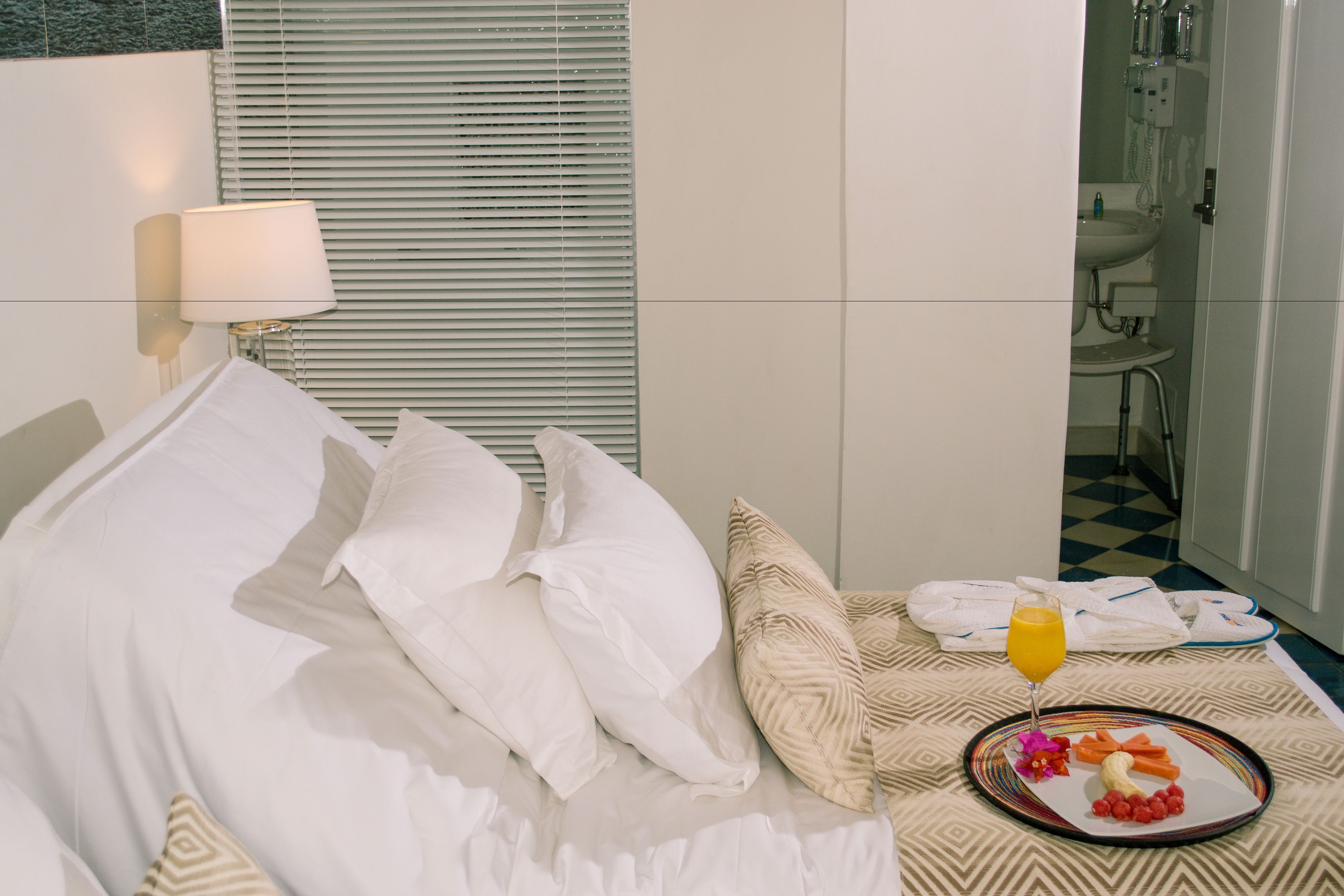 A hotel room with a neatly made bed, a bedside table with a lamp, a tray with food and a glass of orange juice, and a bathroom visible in the background.