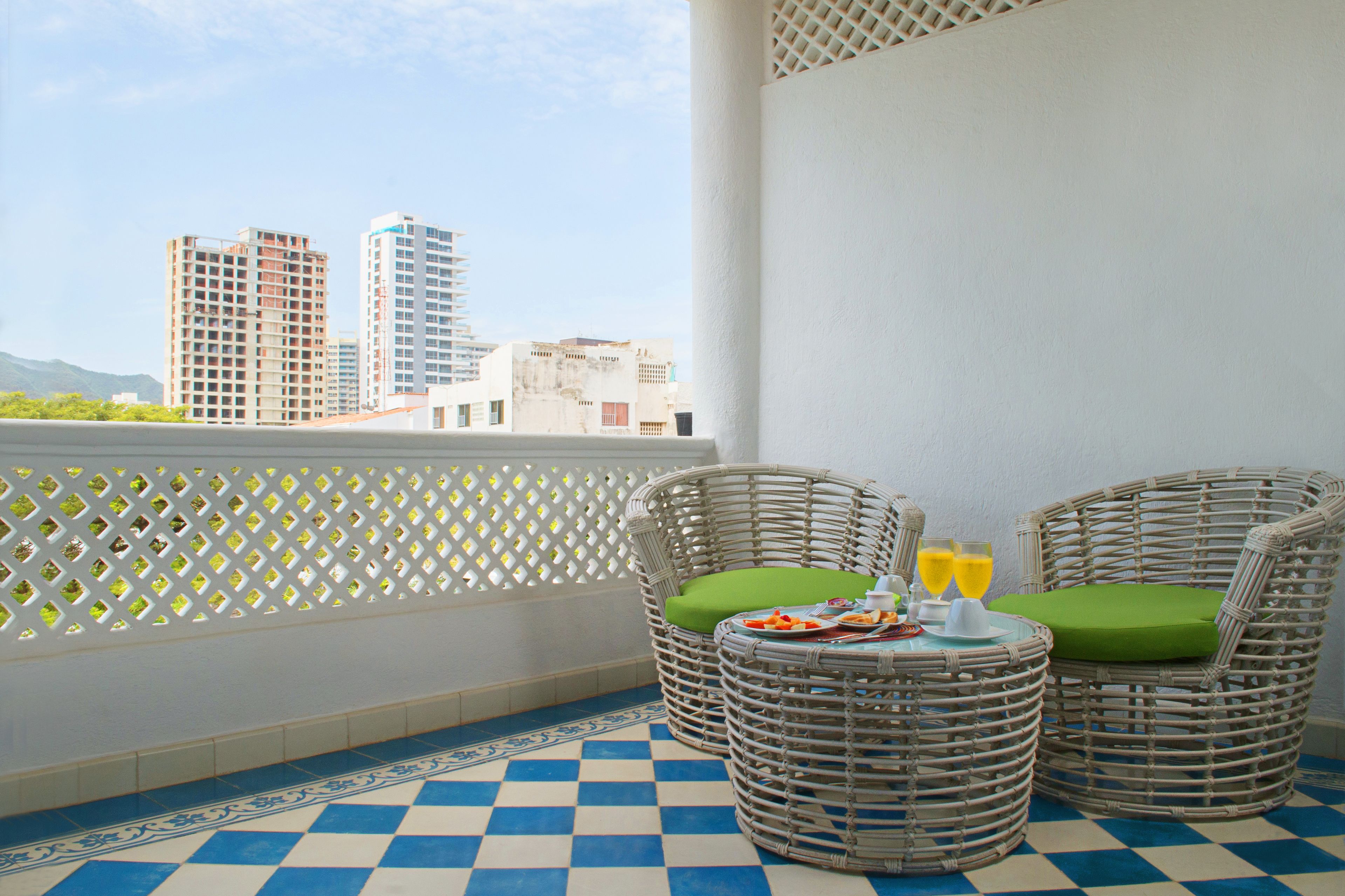 Een balkon met rieten stoelen, een klein tafeltje met drankjes en uitzicht op hoge gebouwen.