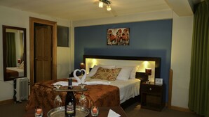 Una habitación de hotel con una cama, una mesita de noche, un radiador, un cuadro colgado en la pared y una mesa con vino, copas y velas.