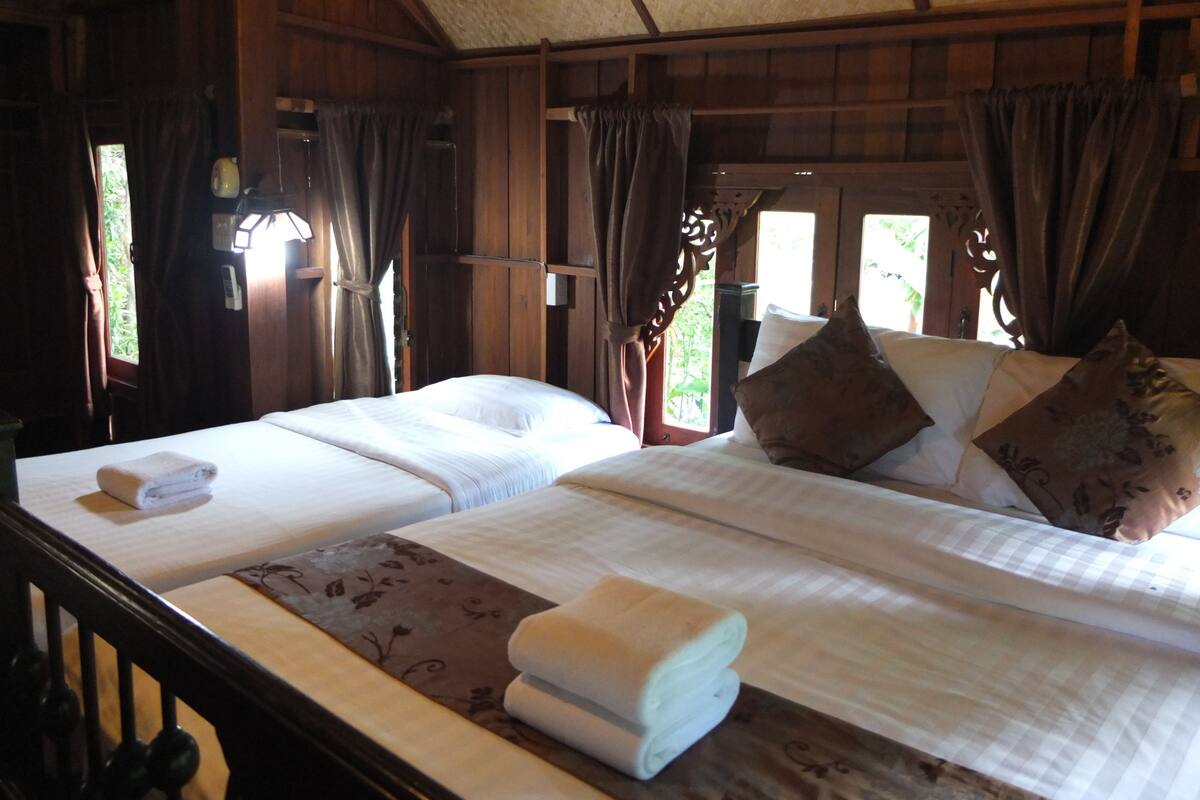 Cama de casal com edredom branco e duas almofadas, cabeceira de madeira e janela com cortinas.