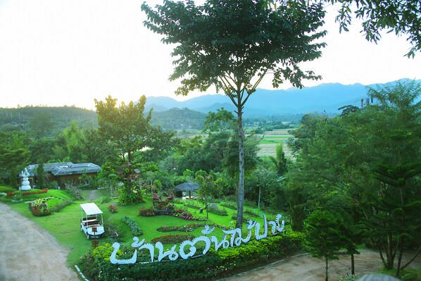 A lush garden with a white car parked in the foreground, surrounded by greenery and a large tree. - Pai Treehouse Resort (Pai)