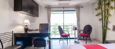 A hotel room with a bed, a desk with a lamp, two chairs, a window with curtains, and a plant.