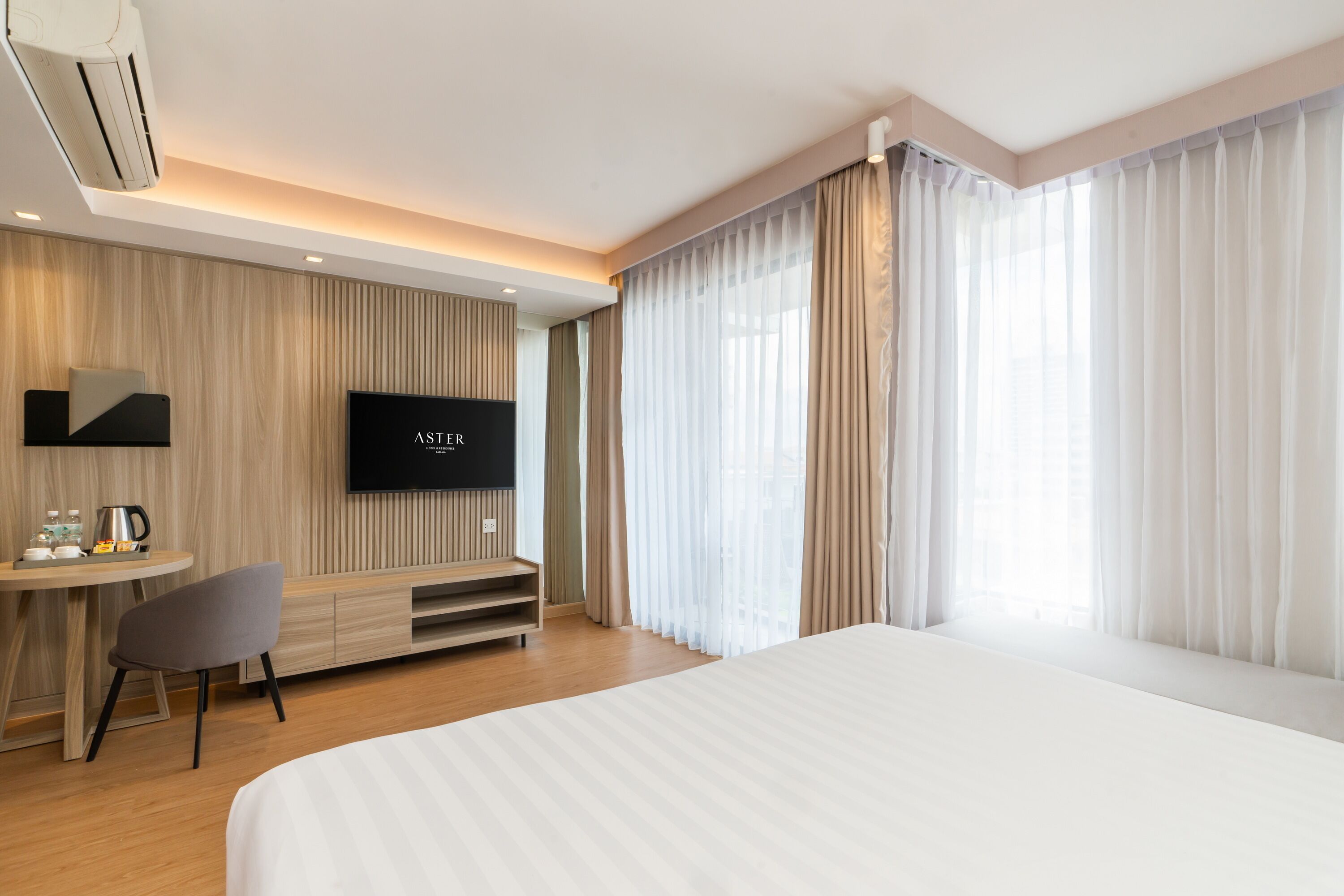 A modern hotel room with a large bed, a TV mounted on the wall, a small desk with a chair, and a window with sheer curtains.
