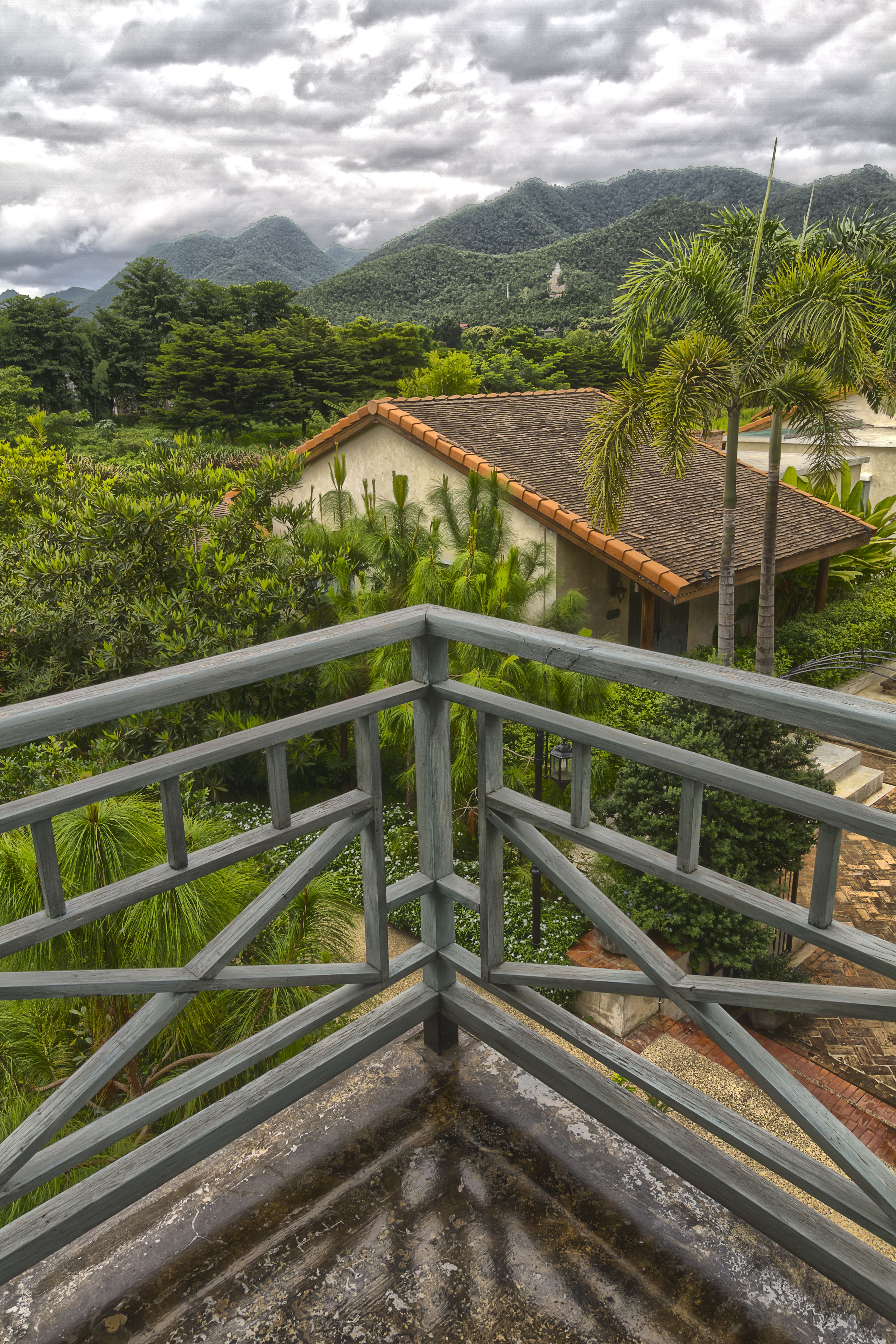 A view from a balcony overlooking a lush garden with a house in the background.
