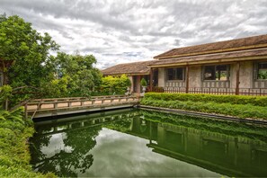 A traditional building with a tiled roof, a bridge over a calm water body, and lush greenery surrounding the area.