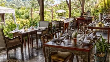 Un restaurant où les tables sont dressées pour le dîner, entouré d’une végétation luxuriante et avec une chaîne de montagnes en toile de fond.