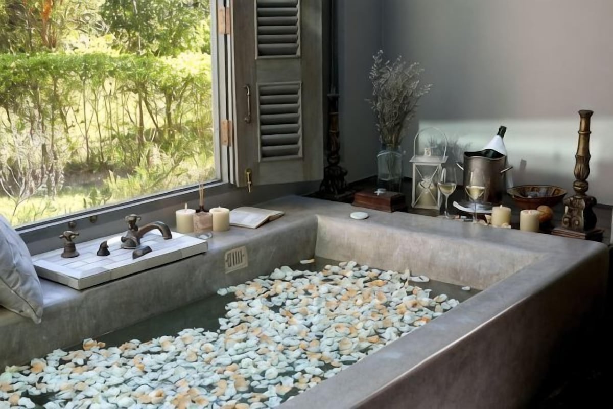 A bathroom with a bathtub filled with pebbles, a window with a view of greenery, and a small table with candles and a tea set.