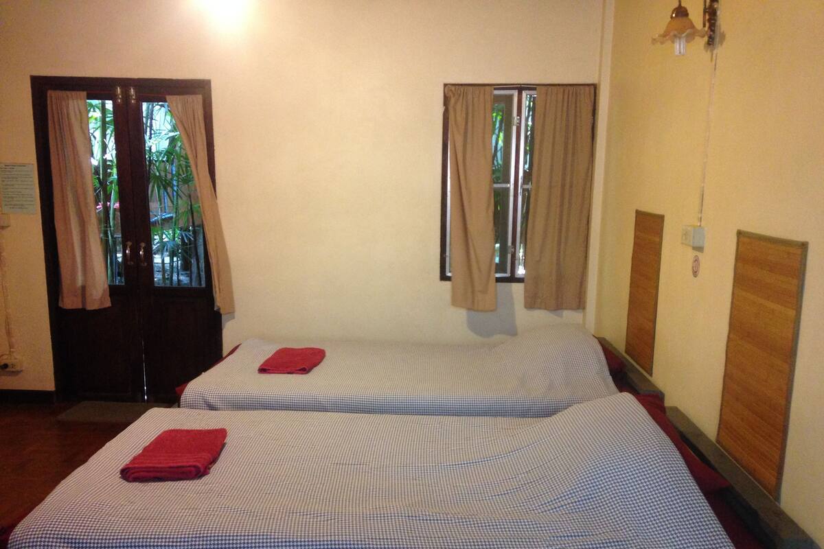 A room with two beds, each with a checkered bedspread and a red towel. There are curtains on the windows, and a light fixture is visible on the wall.