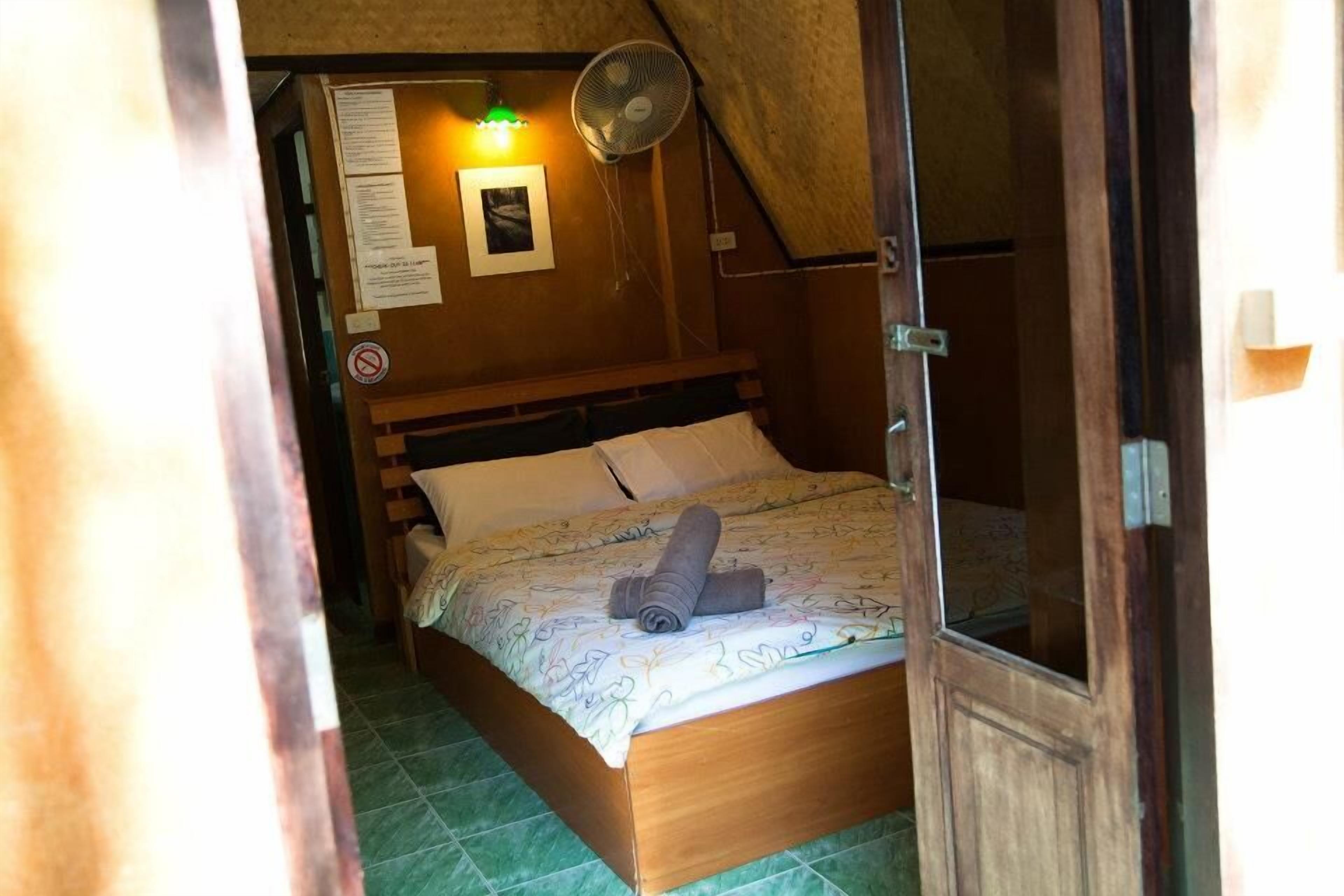 Une petite pièce agréable, avec un lit en bois, un ventilateur fixé au mur et une porte dotée d’un panneau en verre.