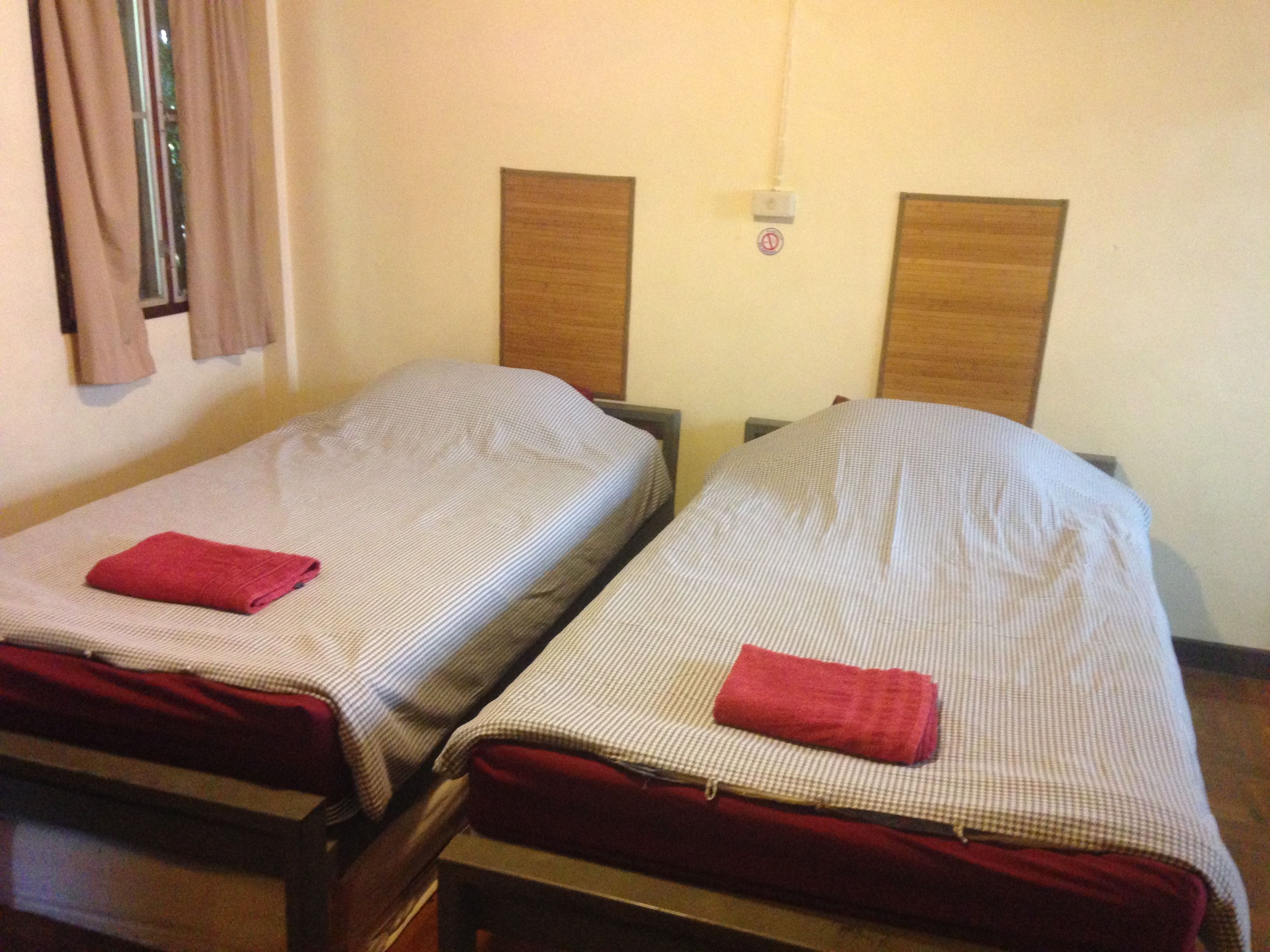 Duas camas de solteiro com roupa de cama xadrez e toalhas vermelhas, cabeceiras de madeira e uma janela com cortinas.