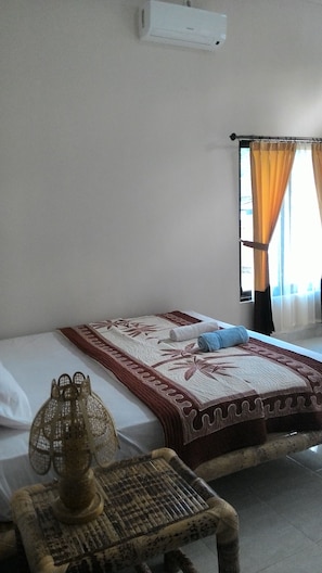 A room with a bed, a small table with a lamp, a window with curtains, and a wall-mounted air conditioner.