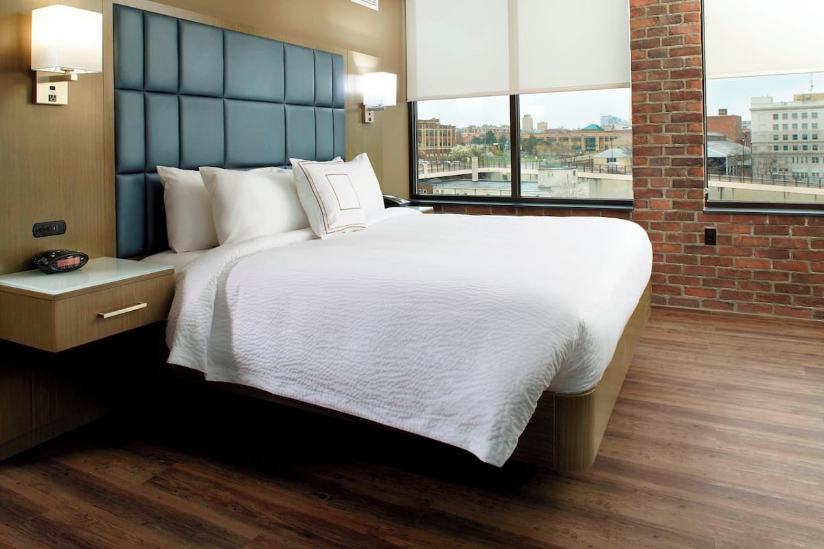 A hotel room with a large bed, wooden flooring, a brick wall, and a city view through the window.