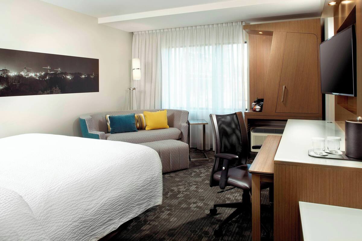 A hotel room with a large bed, a sofa, a desk with a chair, a television, and a window with curtains.