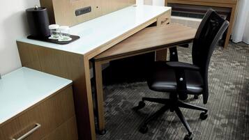 A hotel room with a wooden desk, a black office chair, a small coffee maker, and a pair of glasses on the desk.