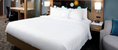 A hotel room with a large bed, a desk, a chair, and a nightstand with a lamp.