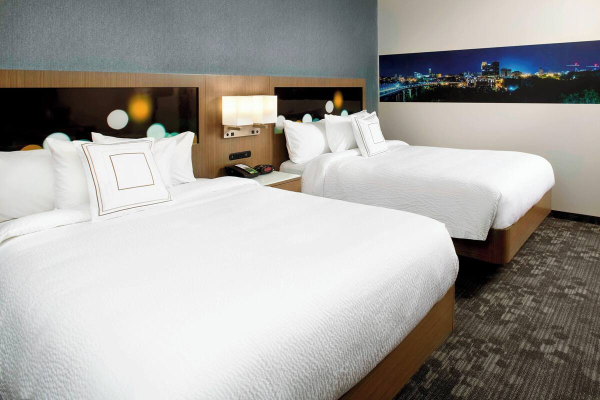 A hotel room with two beds, a nightstand, a lamp, and a cityscape view.
