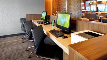 Zona de recepción de hotel con dos estaciones de trabajo de computadora, un mostrador de recepción y un área de estar con mesas y sillas.
