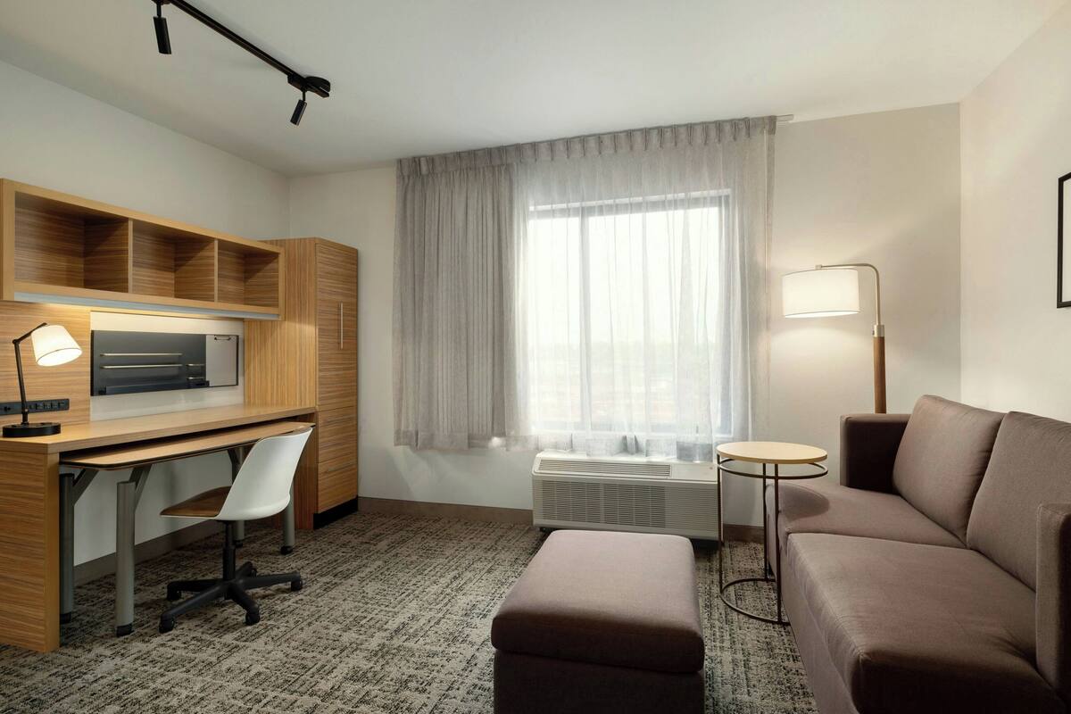 Une chambre d’hôtel avec un bureau en bois, une chaise blanche, un canapé marron, une petite table ronde, un lampadaire et une grande fenêtre avec des rideaux légers.