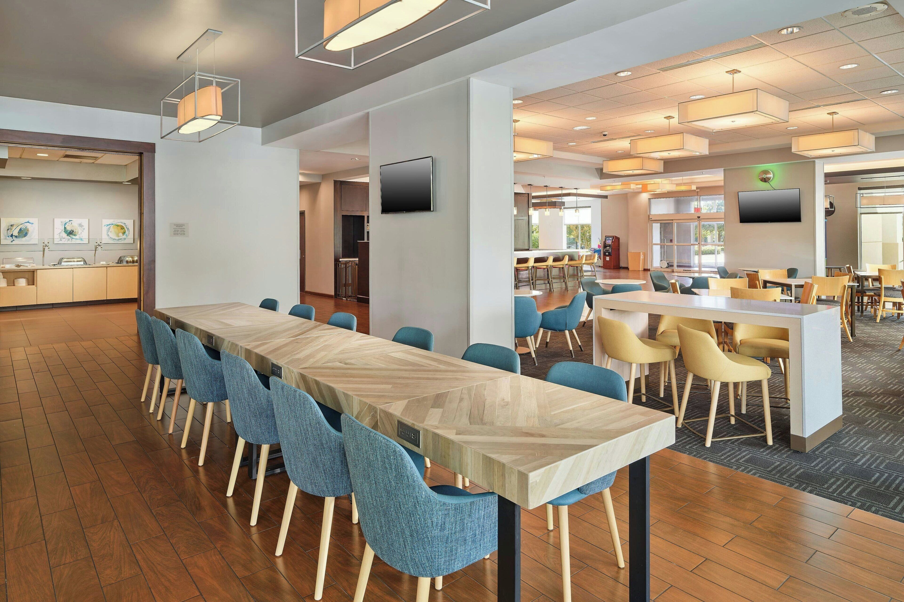 A modern dining area with a long table and blue chairs, a bar area with stools, and a kitchen in the background.