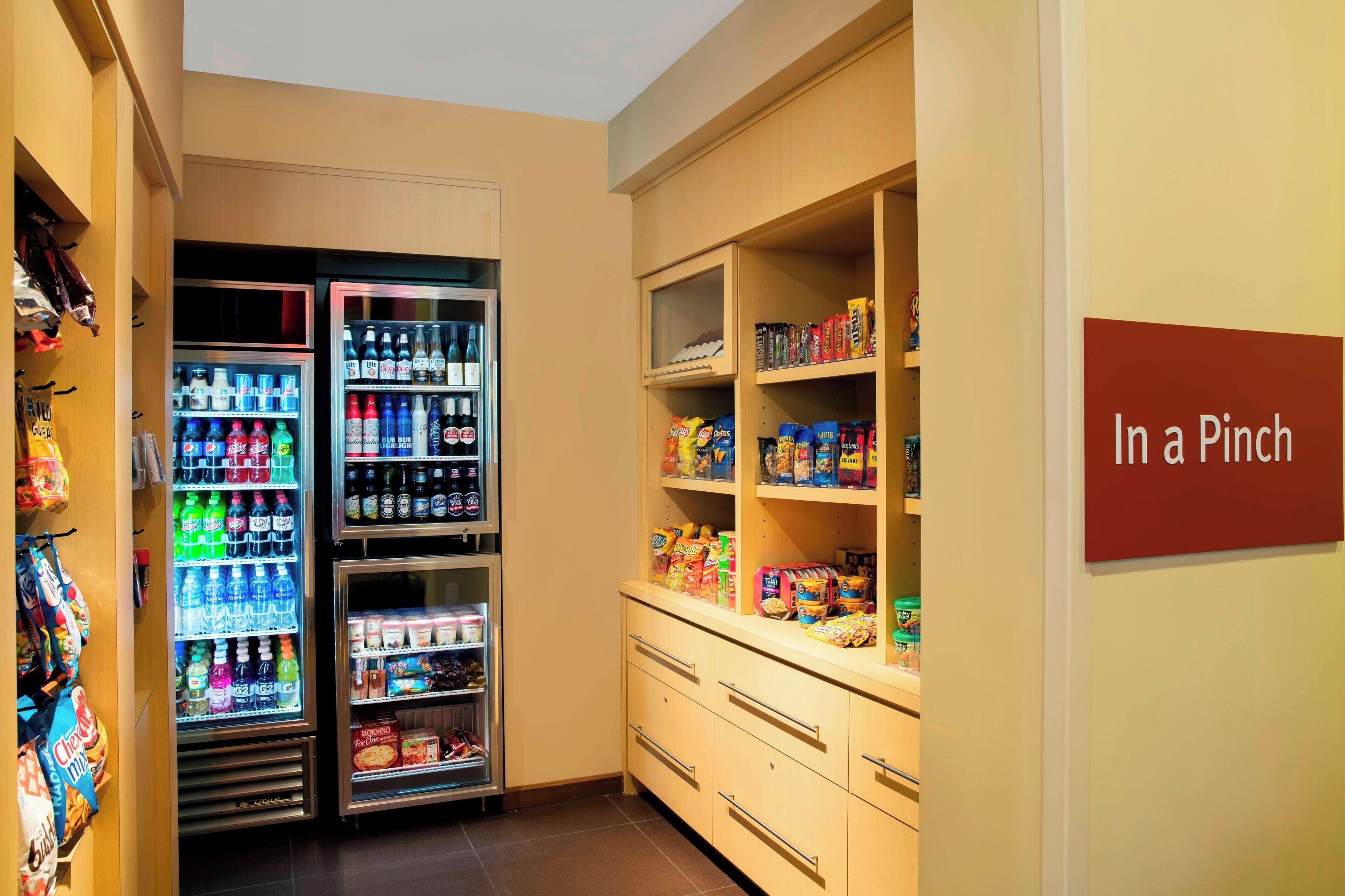 A hotel pantry with refrigerated beverage dispensers, snack shelves, and a sign reading "In a Pinch".