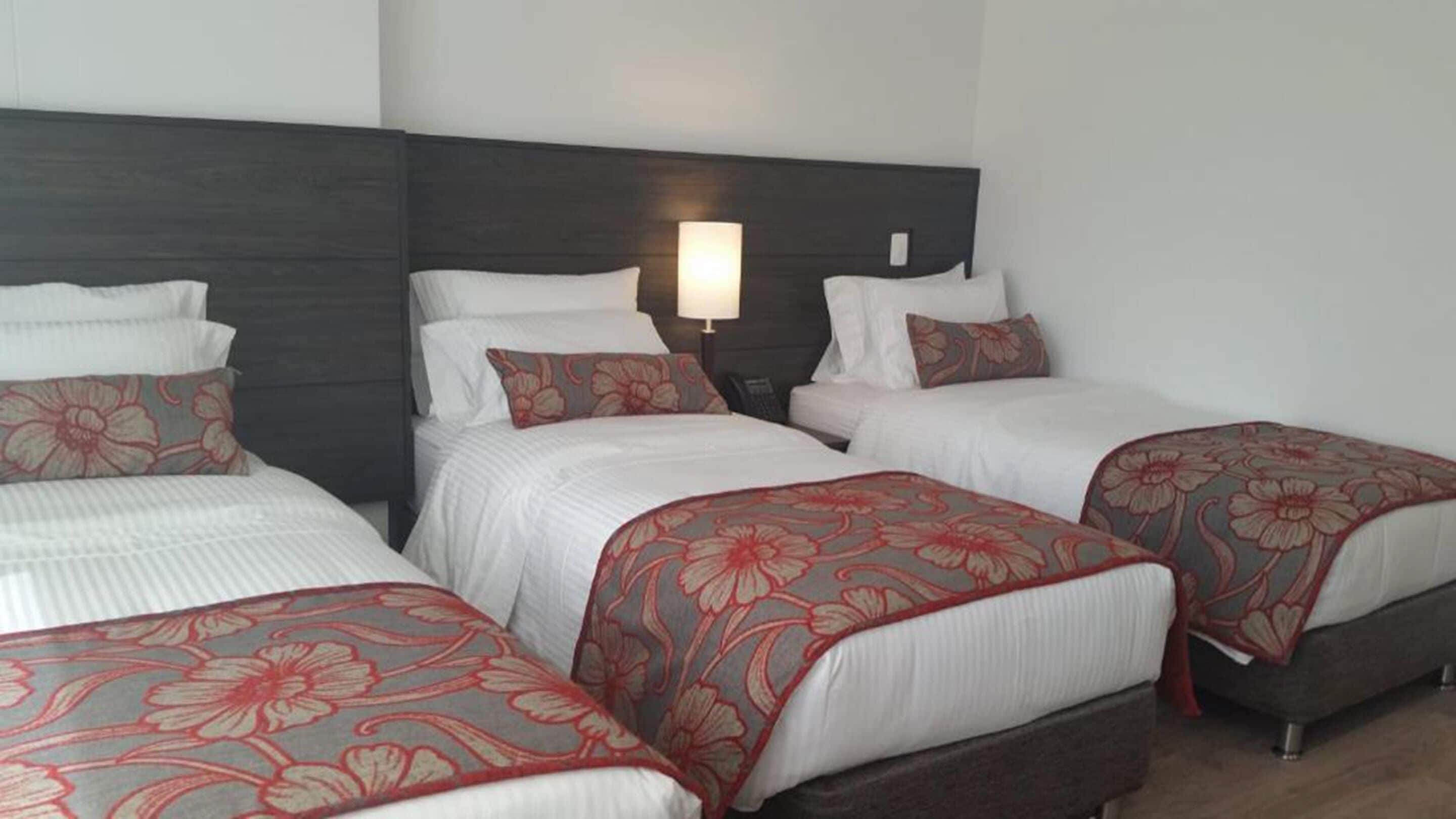 Quarto de hotel com três camas de solteiro, cada uma com colcha estampada em flor e almofadas brancas. Uma luminária de cabeceira é visível em uma das camas.